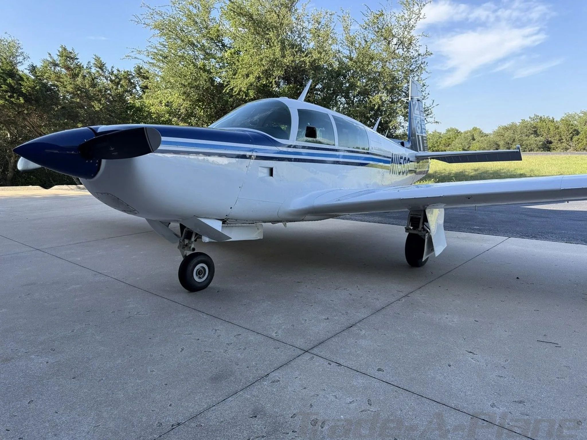 A front-side view of the white and blue single-engine aircraft, N1156G, parked on concrete with trees in the background.