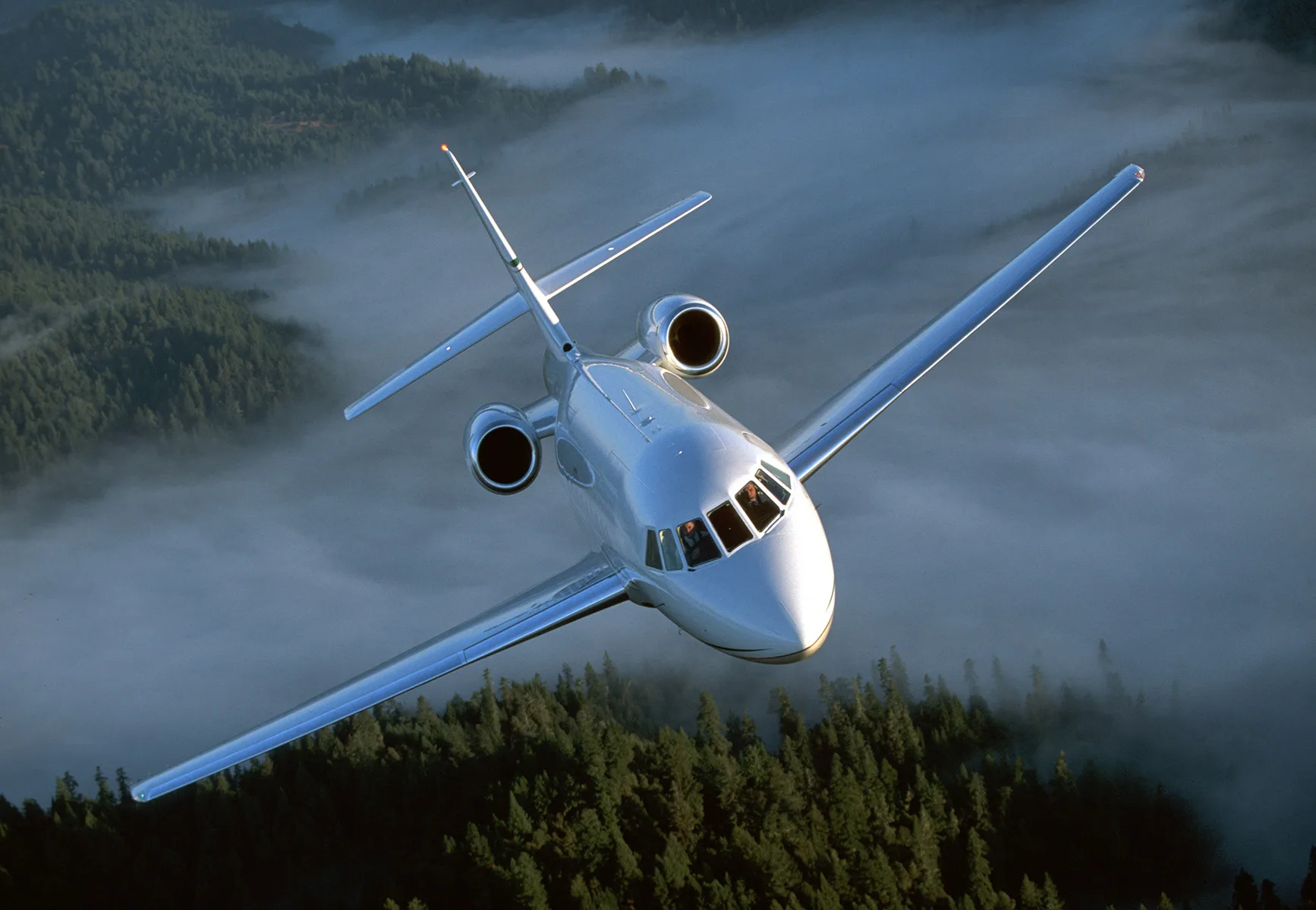 A white business jet with two rear-mounted engines flies over a misty, forested landscape.