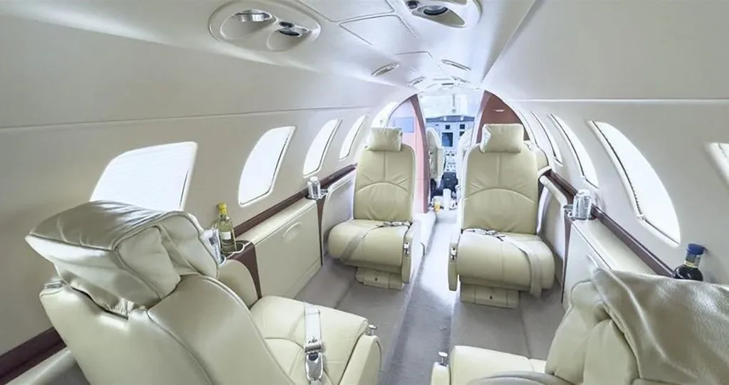 A wide-angle view of a private jet's luxurious light beige leather cabin interior, looking towards the cockpit.