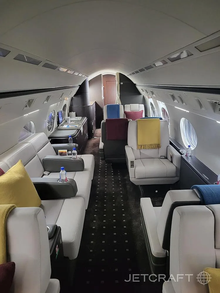 The spacious interior of a private jet, featuring a mix of individual seats and a long sofa, all in light colors with colorful accents.