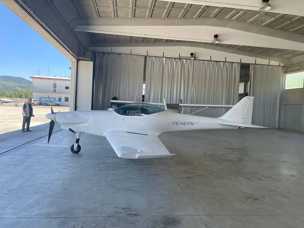 A white Fascination ultralight aircraft is partially outside a hangar with a person standing nearby, viewed from the side.