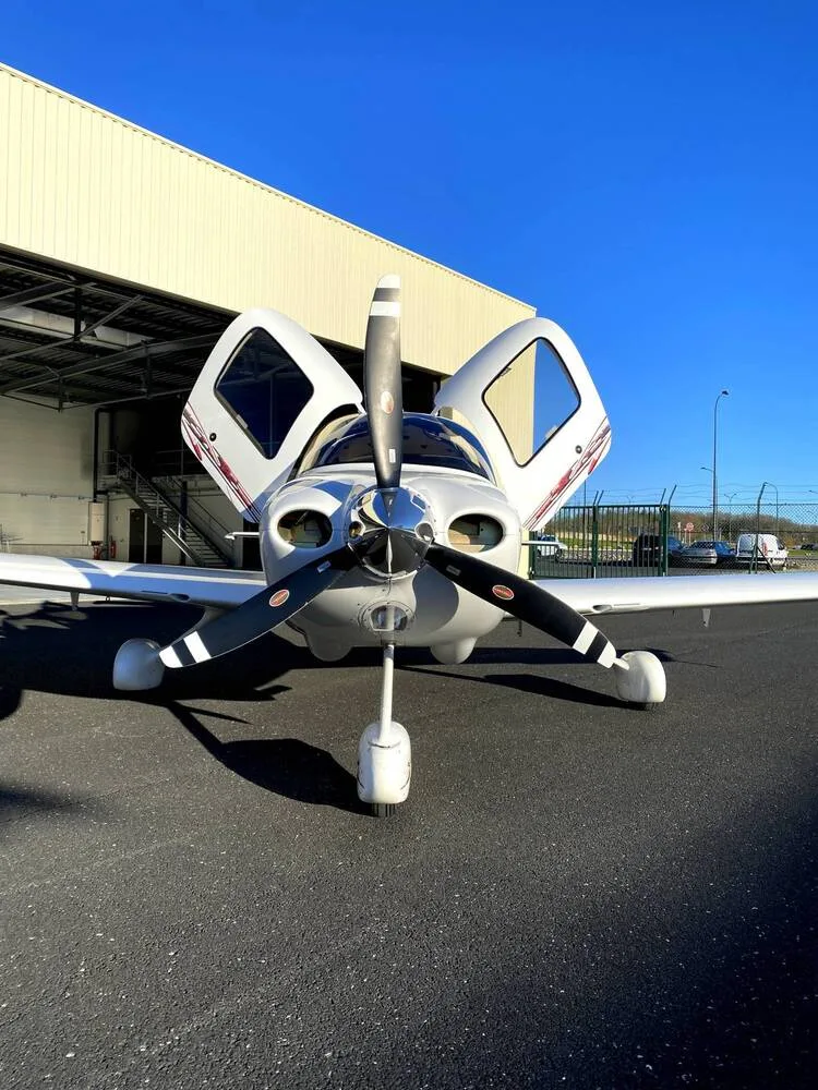 A head-on view of a white Cirrus SR22 aircraft with its gull-wing doors open, parked on an airfield.