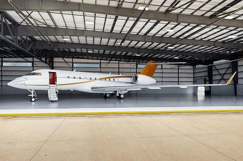 A white and gold private jet, likely a Bombardier Challenger, is parked inside a hangar with its main door open and stairs extended.