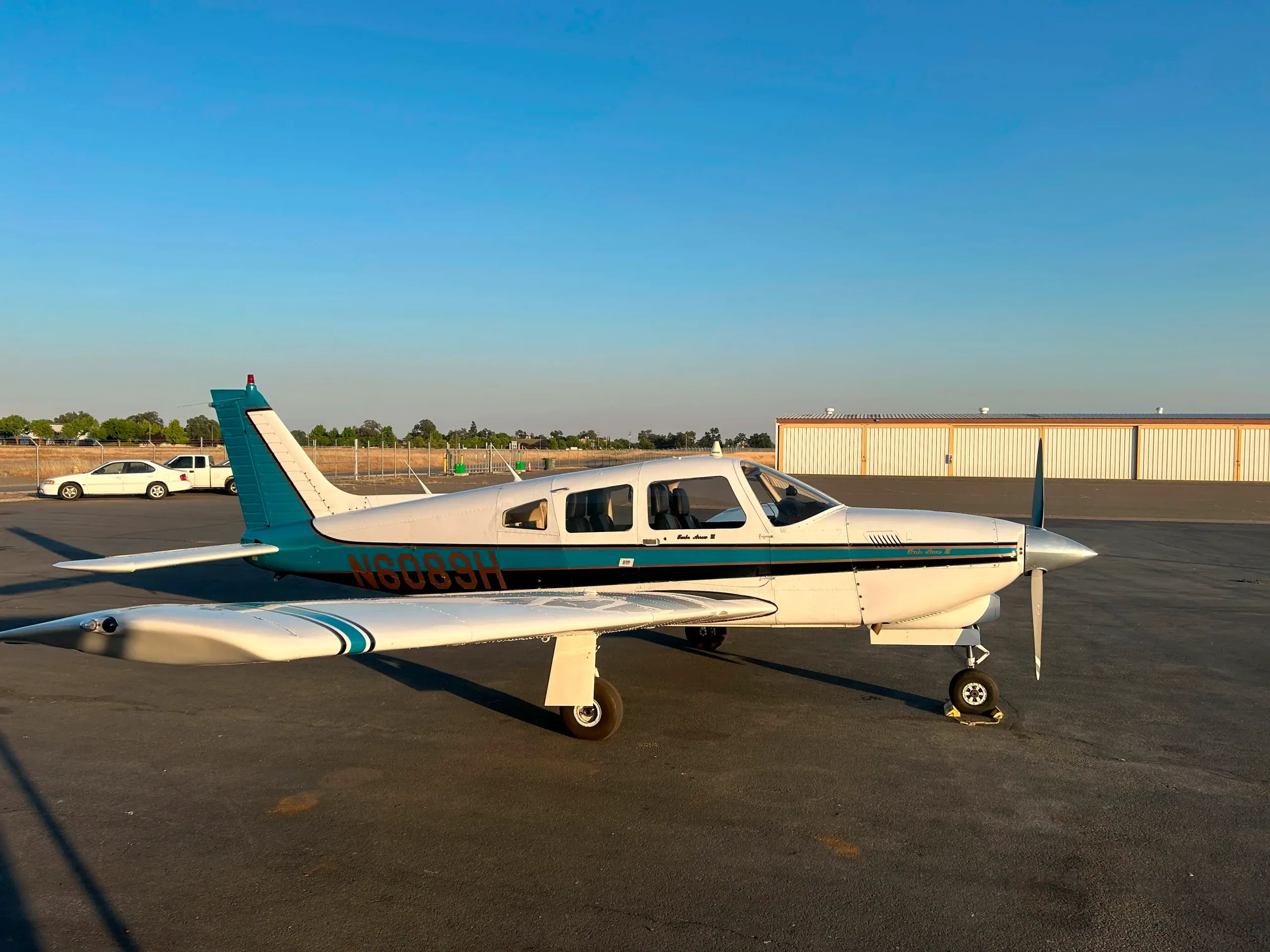 A white and teal single-engine propeller aircraft, N6089H, is parked on an asphalt tarmac under a clear blue sky.