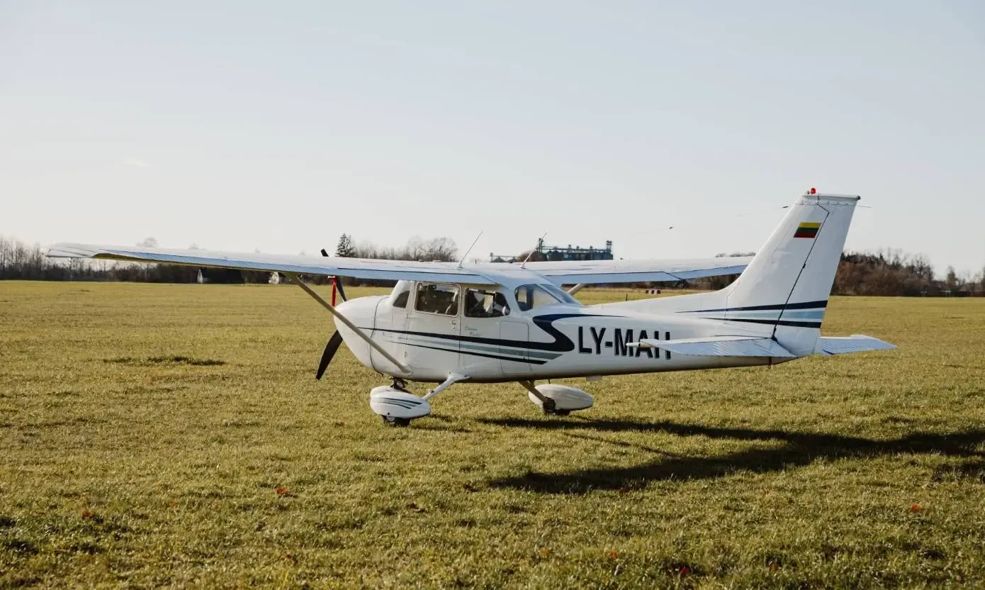 A white Cessna 172 with registration LY-MAH and a Lithuanian flag on its tail, parked on a sunny grassy field.