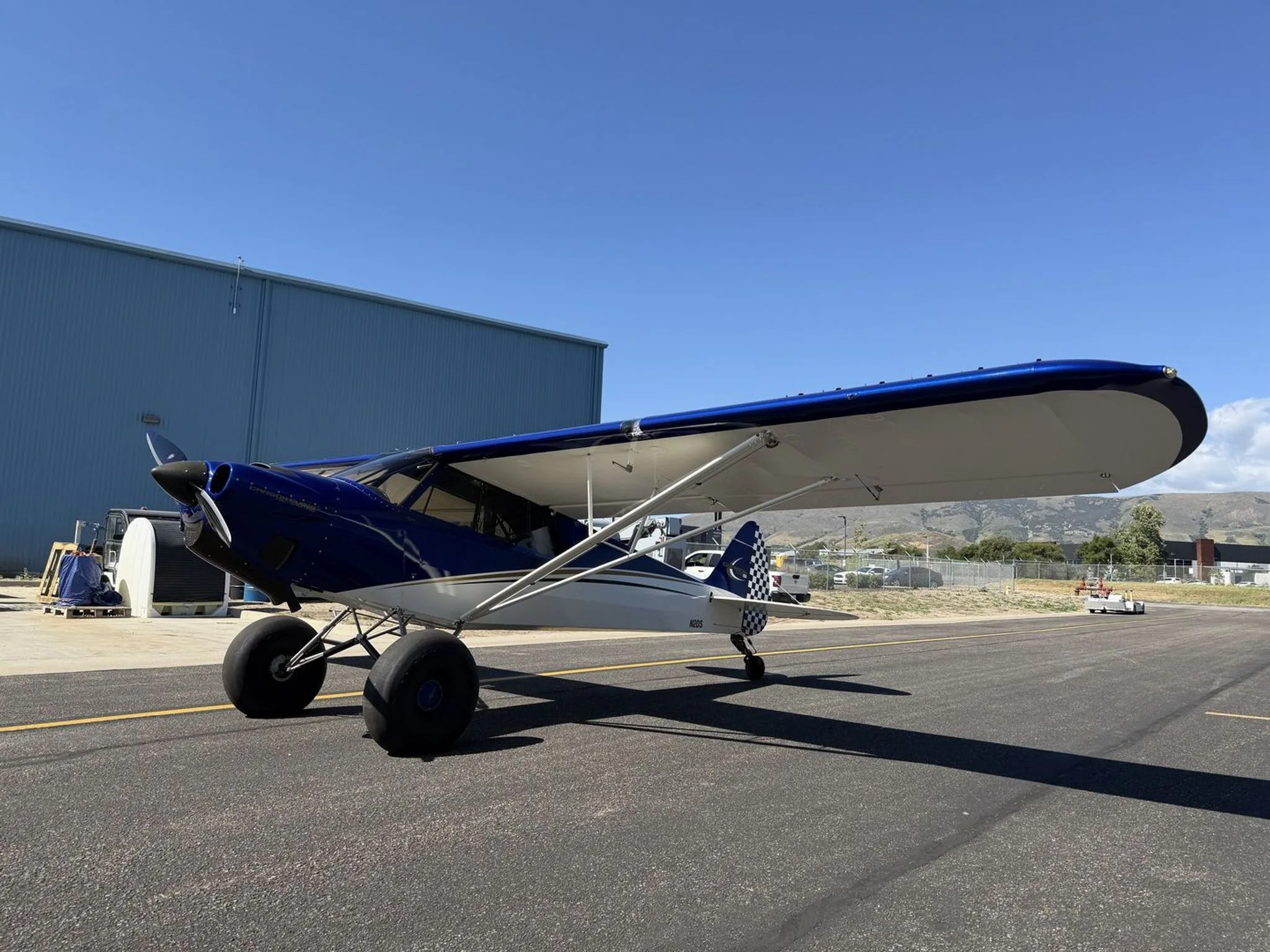 A blue and white Carbon Cub SS aircraft with large tundra tires, seen from the left side on an airport tarmac.