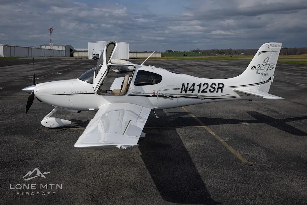 A white Cirrus SR22T aircraft with open doors is parked on an asphalt runway with hangars in the background.