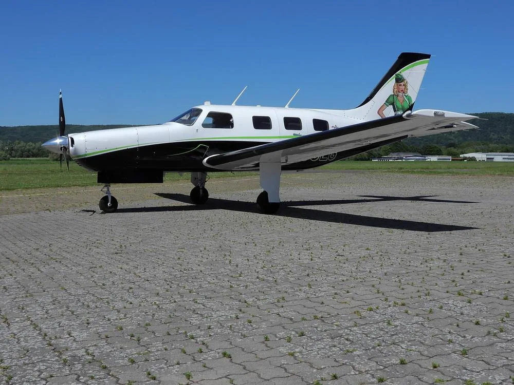 A side view of a white and black Piper M-Class aircraft with green accents and a pin-up girl on the tail, parked on a paved surface.