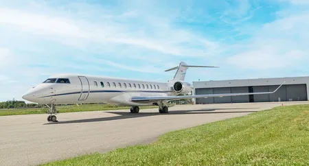 A white business jet with blue accent stripes is parked on an asphalt tarmac next to a hangar under a partly cloudy sky.