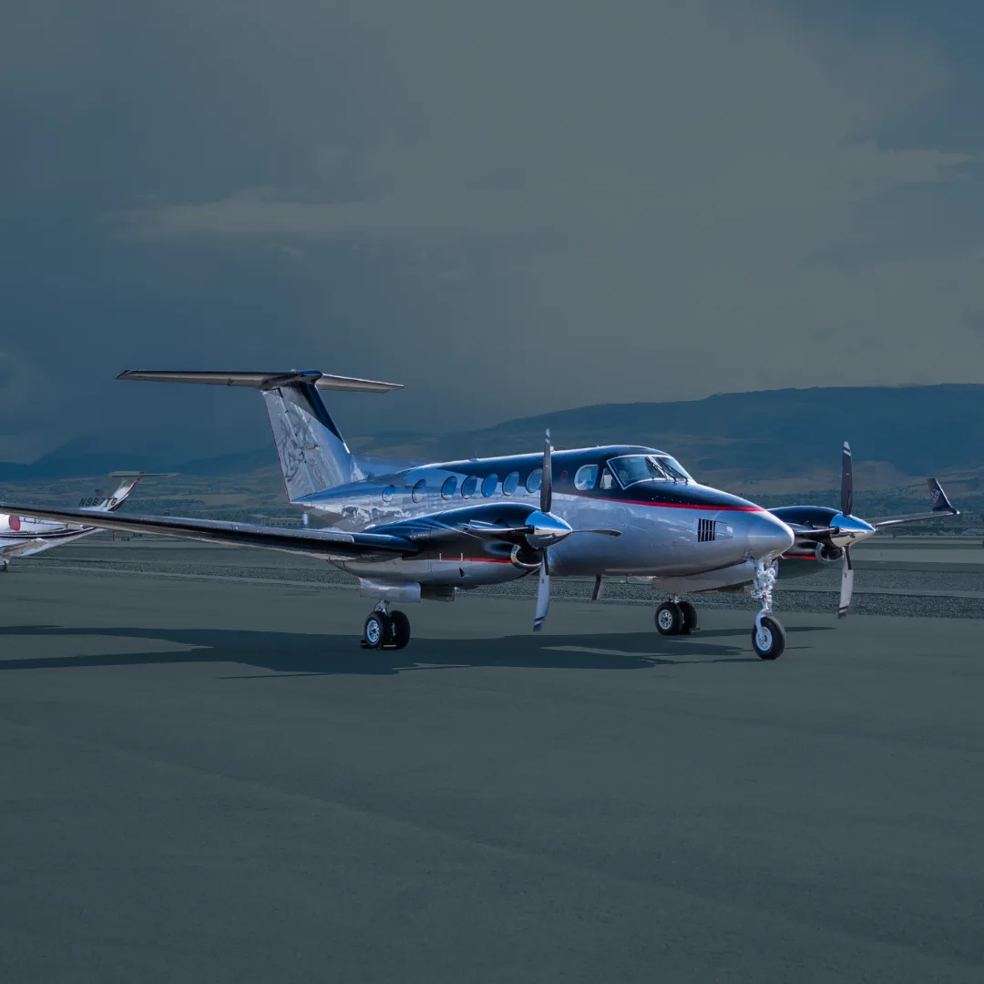 A silver and black twin-engine turboprop aircraft, possibly a King Air, parked on an airfield with mountains in the background.
