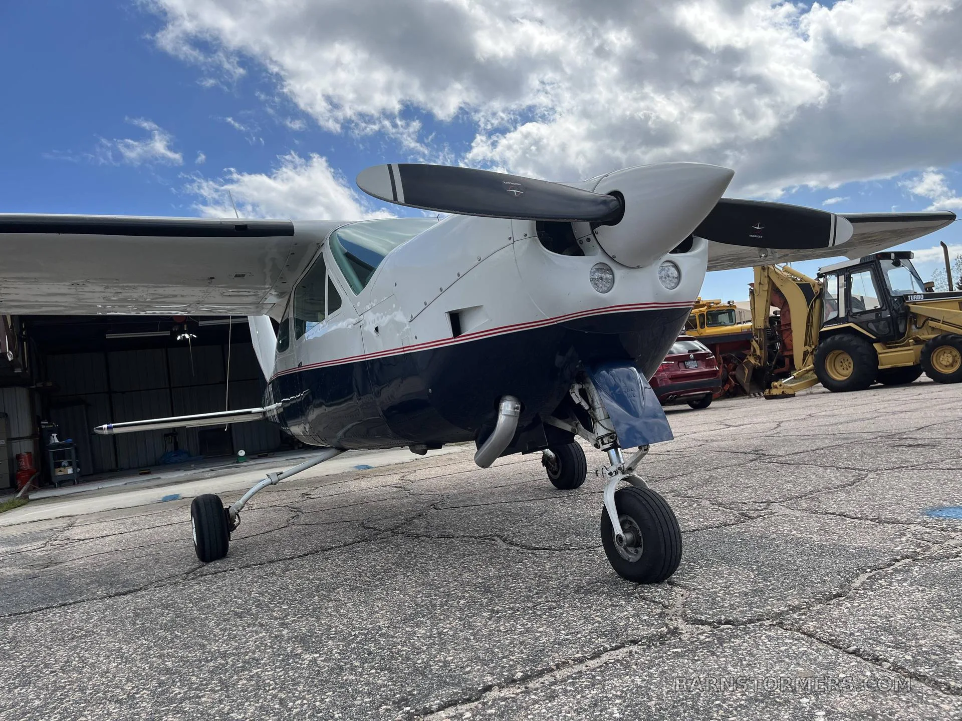 Close-up, low-angle front view of the propeller and nose of the white and dark blue single-engine aircraft N1217M, with construction equipment in the background.