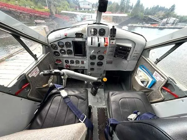 The cockpit of a floatplane with a detailed instrument panel, two pilot seats, and a view of the water outside.