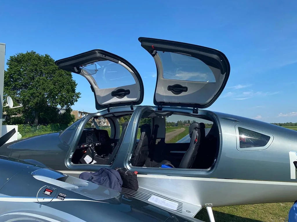 A close-up side view of the open gull-wing doors of a dark grey Diamond DA62 aircraft, revealing the cockpit and cabin seats.