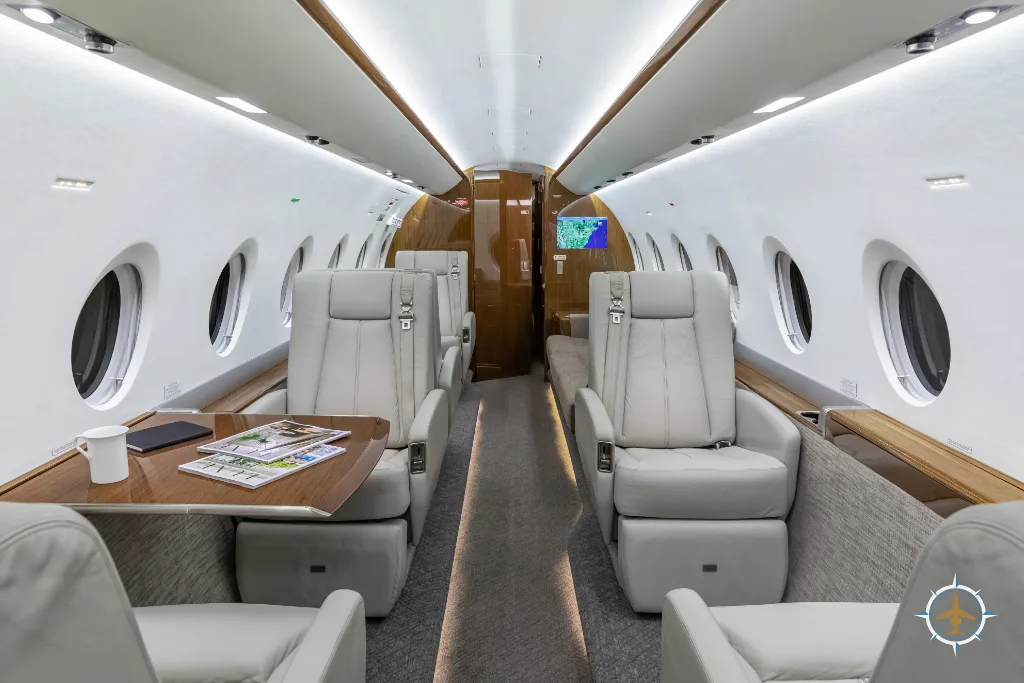 The luxurious aircraft cabin with light grey leather seats, a wooden table, and large windows.
