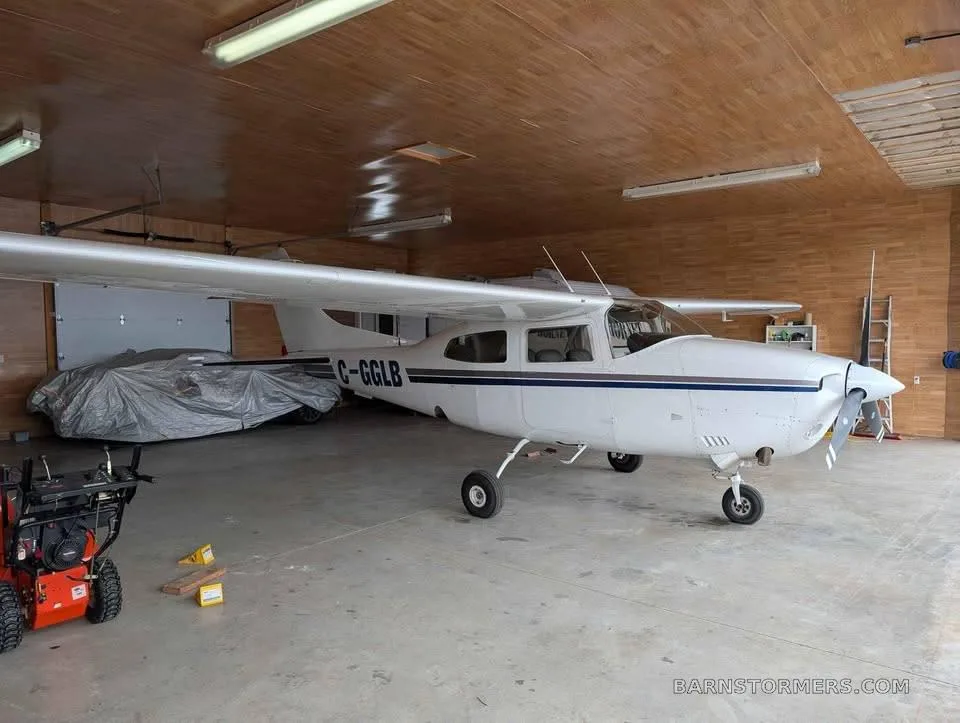 A white Cessna 182 with blue stripes, registration C-GGLB, parked in a hangar.
