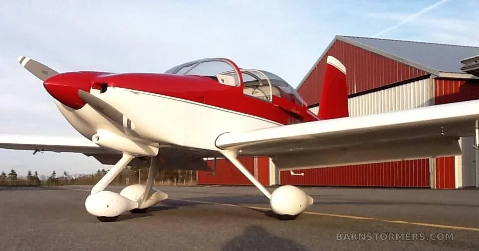 A red and white single-engine experimental aircraft is parked on an asphalt tarmac next to a red hangar.