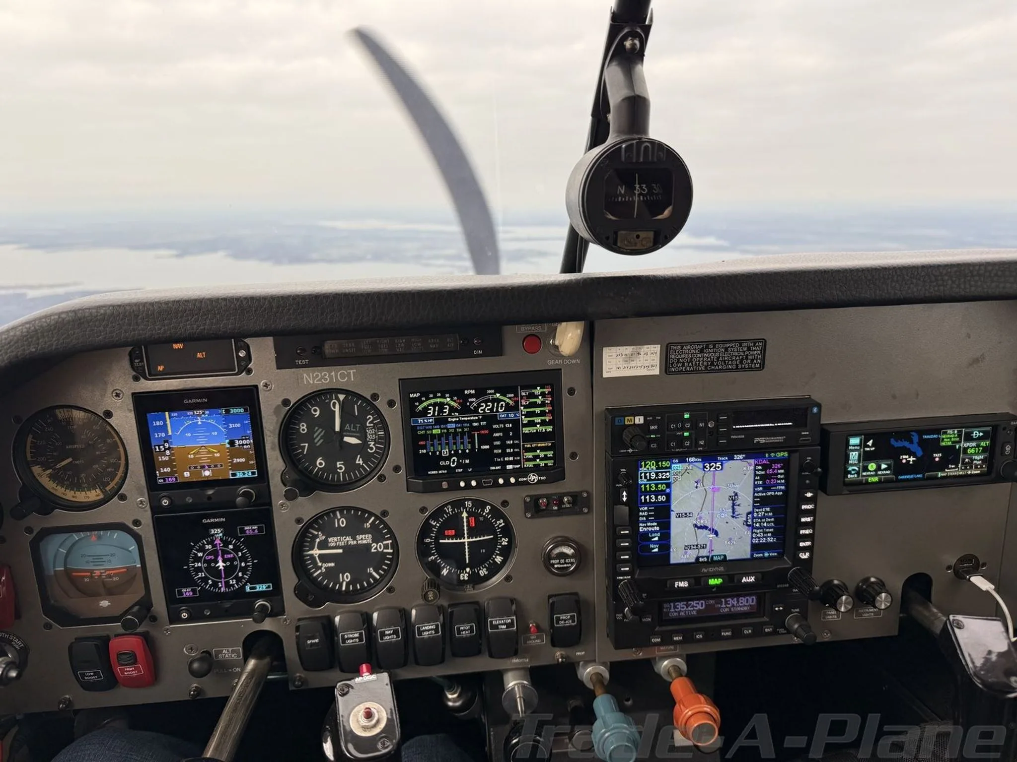 Cockpit view showing the instrument panel with multiple Garmin displays and analog gauges, with a view of water and land outside.