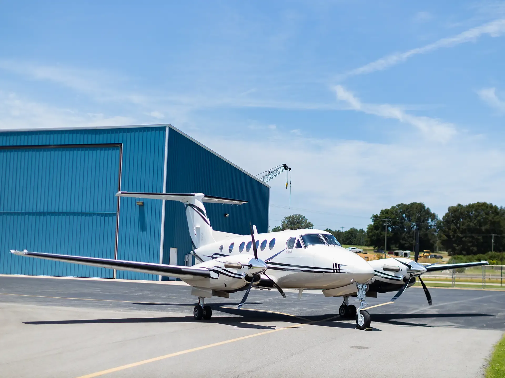 A white Beechcraft King Air twin-turboprop aircraft with black stripes is parked on an asphalt apron in front of a blue hangar under a clear sky.