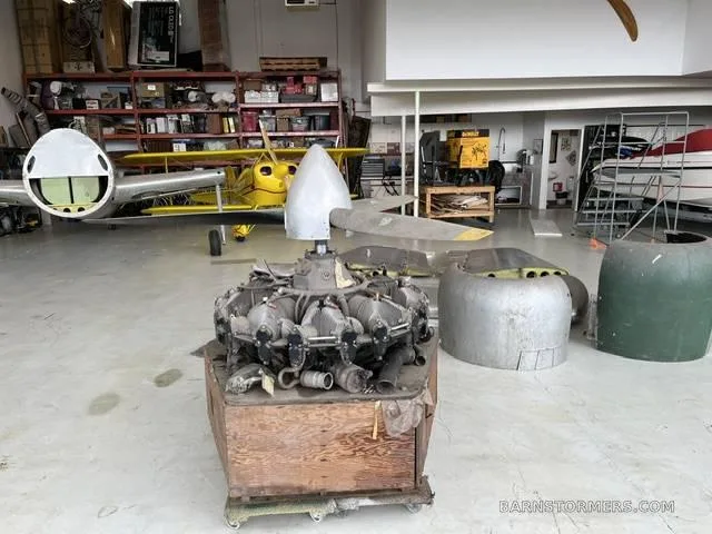 A large radial aircraft engine with a propeller is on a wooden crate inside a hangar, with other aircraft parts and a yellow biplane in the background.