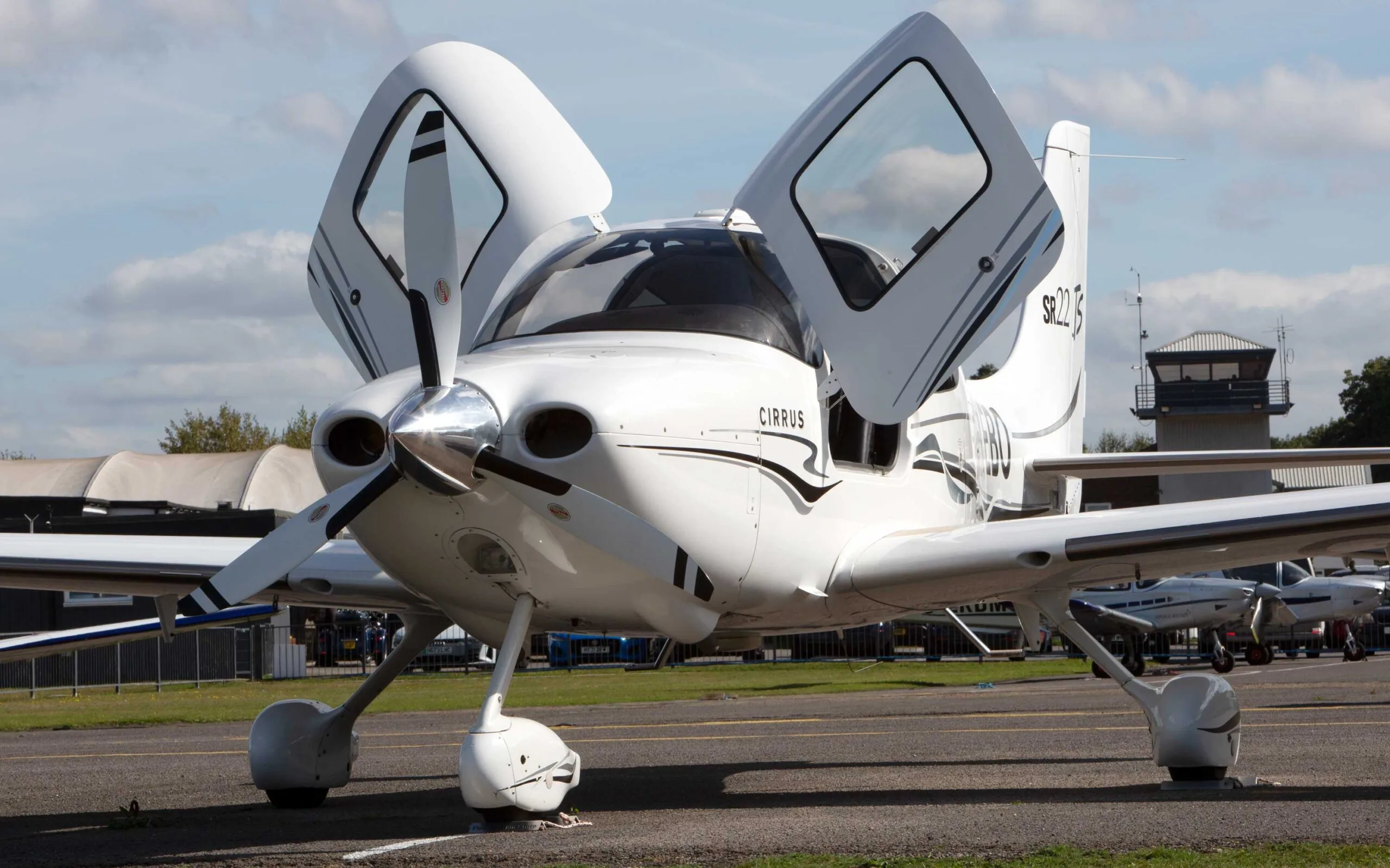 A white Cirrus SR22 TSi aircraft with black and grey stripes, seen from the front-left with its gull-wing doors open on an airfield.