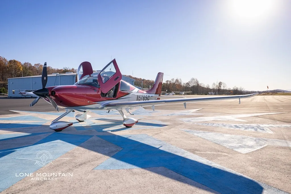A red and white Cirrus SR22 aircraft with registration N5785C parked on an airfield with its doors open, a hangar in the background.