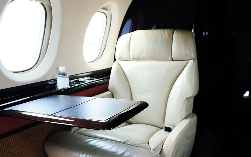 A cream-colored leather aircraft seat with a fold-out wooden table and two oval windows.