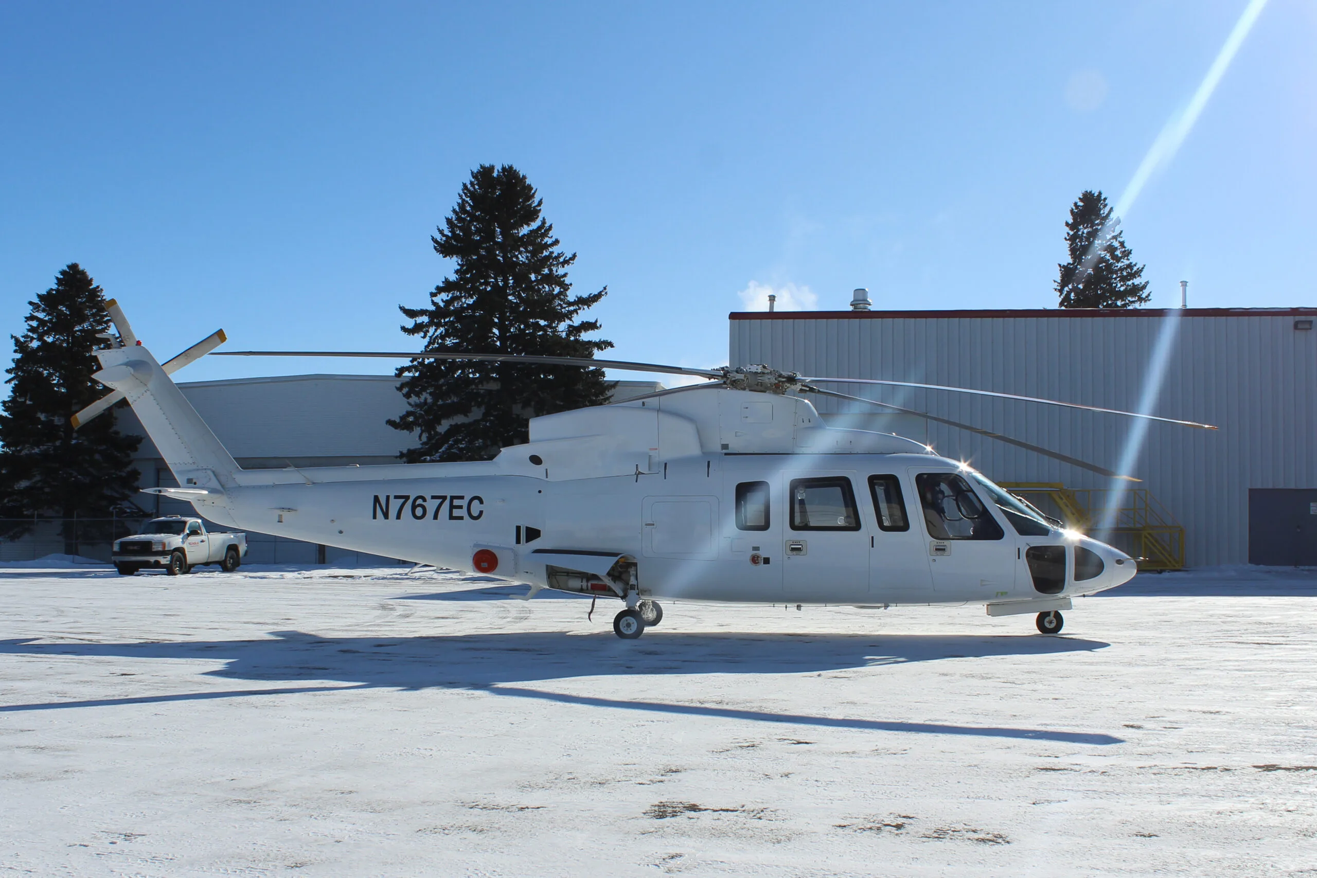 A white AgustaWestland AW139 helicopter with registration N767EC sits on a snowy tarmac in front of a hangar on a sunny day.