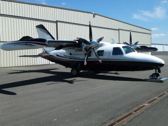 A white and black Mitsubishi MU-2 twin-turboprop aircraft with red and grey stripes, parked on an asphalt ramp in front of a hangar.