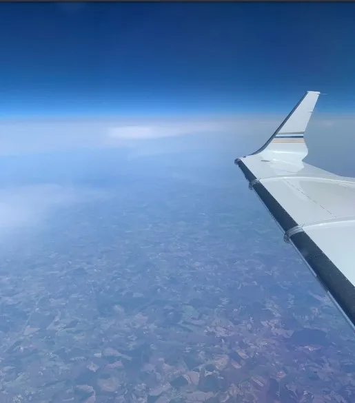 View from an aircraft window showing the white wing with blue and gold stripes above clouds and a patchwork of land.