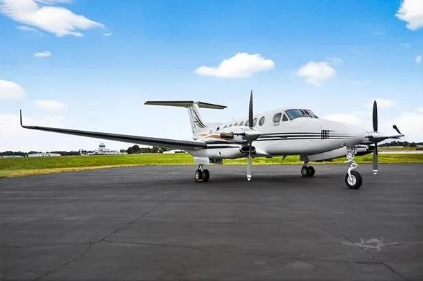 A white Beechcraft King Air with black and gold stripes is parked on an asphalt tarmac under a blue sky.