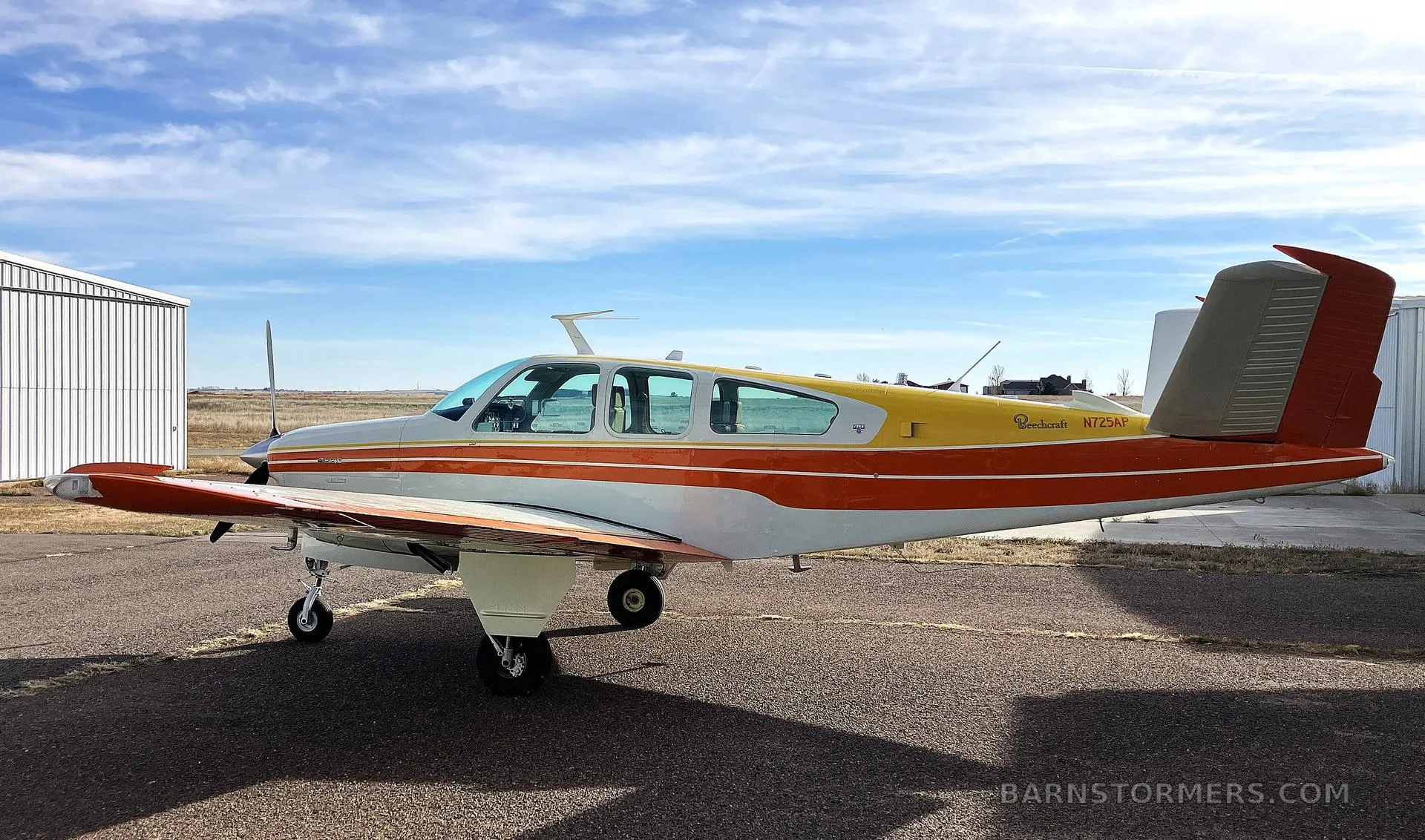 A side view of a white, orange, and yellow Beechcraft Bonanza N725AP parked on an airfield with a hangar in the background.