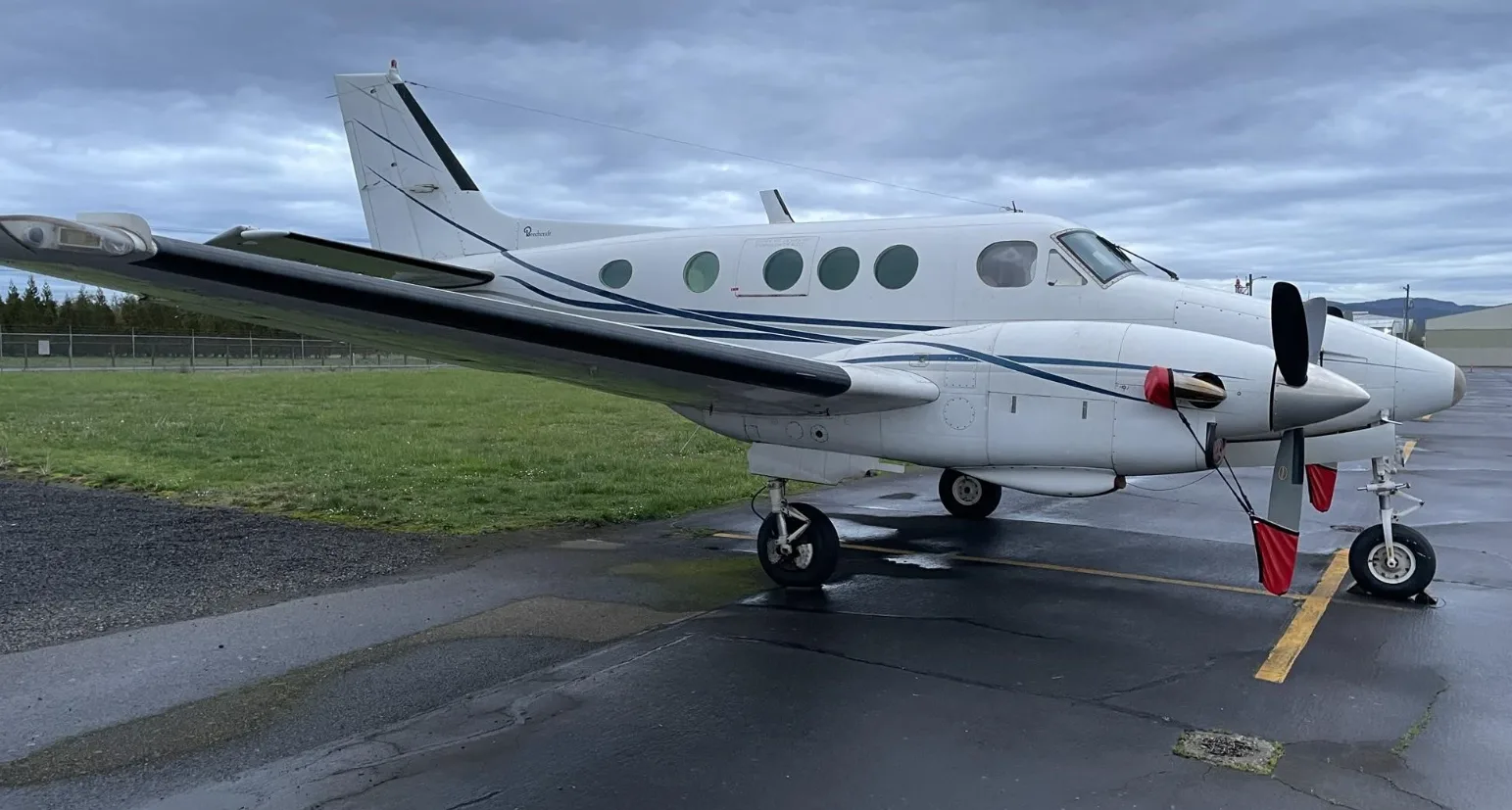 A white Beechcraft King Air 200 with blue stripes is parked on a wet tarmac under a cloudy sky.