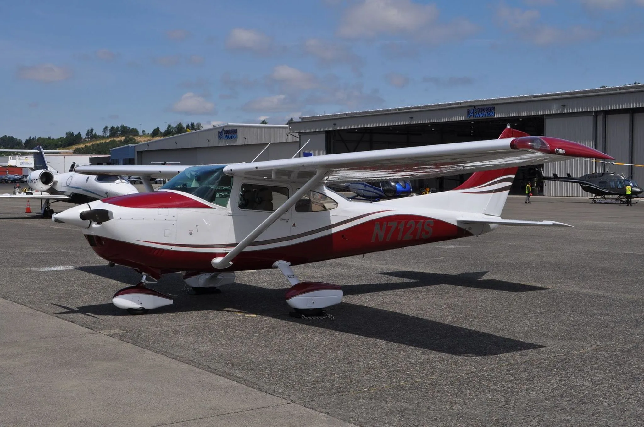 A red and white Cessna N7121S is parked on an airfield in front of hangars.