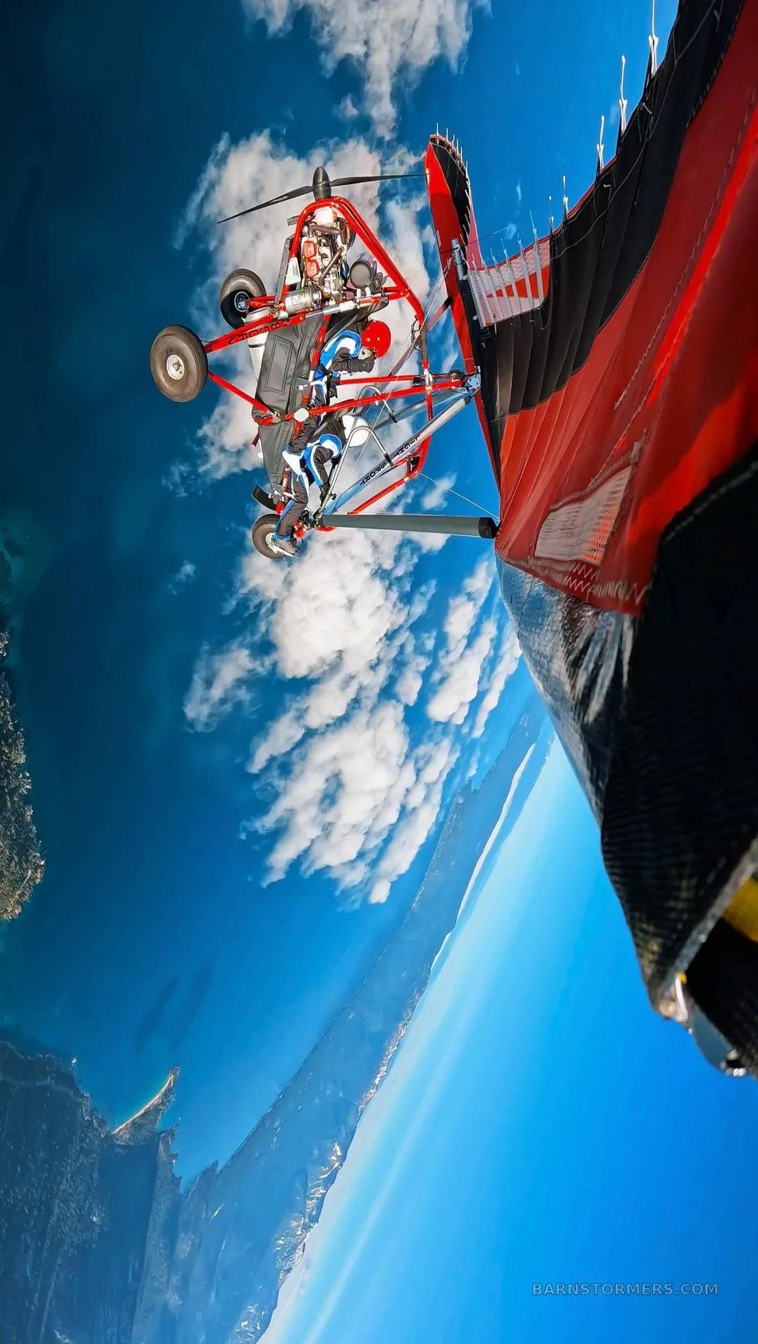 A red and black trike ultralight aircraft in flight, seen from above, with a vast blue sky, clouds, and a mountainous landscape with water below.