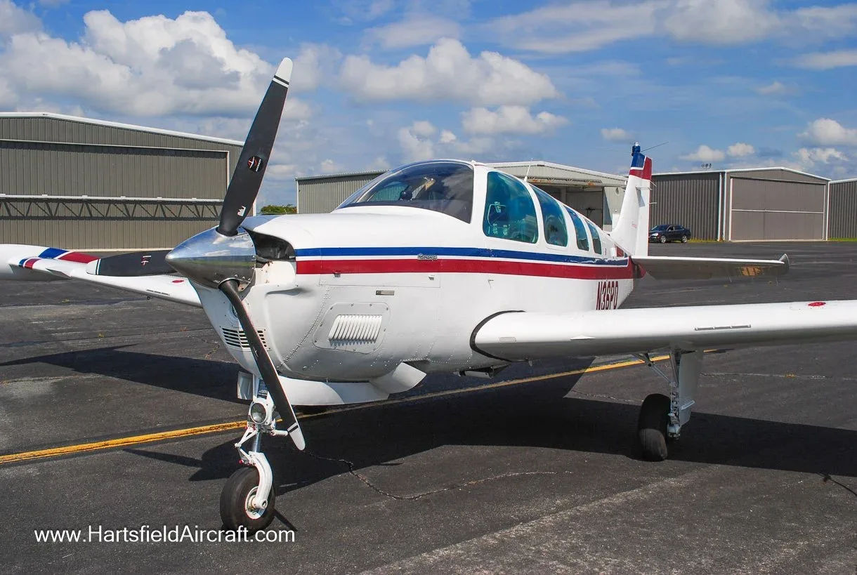 A white Beechcraft Bonanza aircraft with red and blue stripes is parked on an airfield tarmac with hangars in the background under a cloudy sky.
