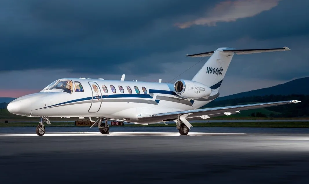 A white Cessna Citation CJ3 business jet (N906MC) with blue and grey stripes is parked on a tarmac at dusk.