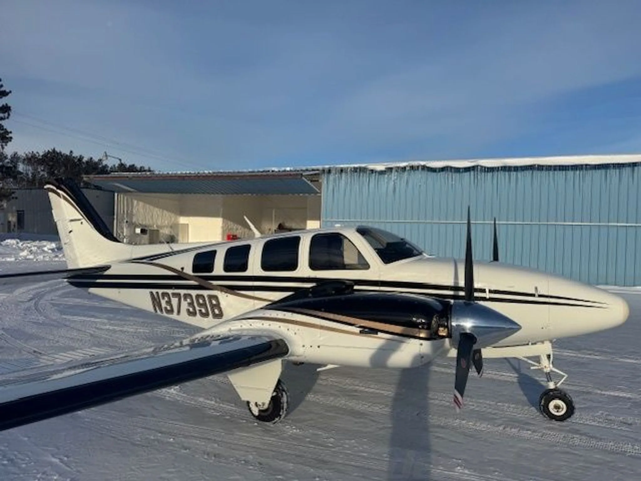 A white and black twin-engine aircraft with gold stripes parked on a snowy ground in front of a blue hangar.