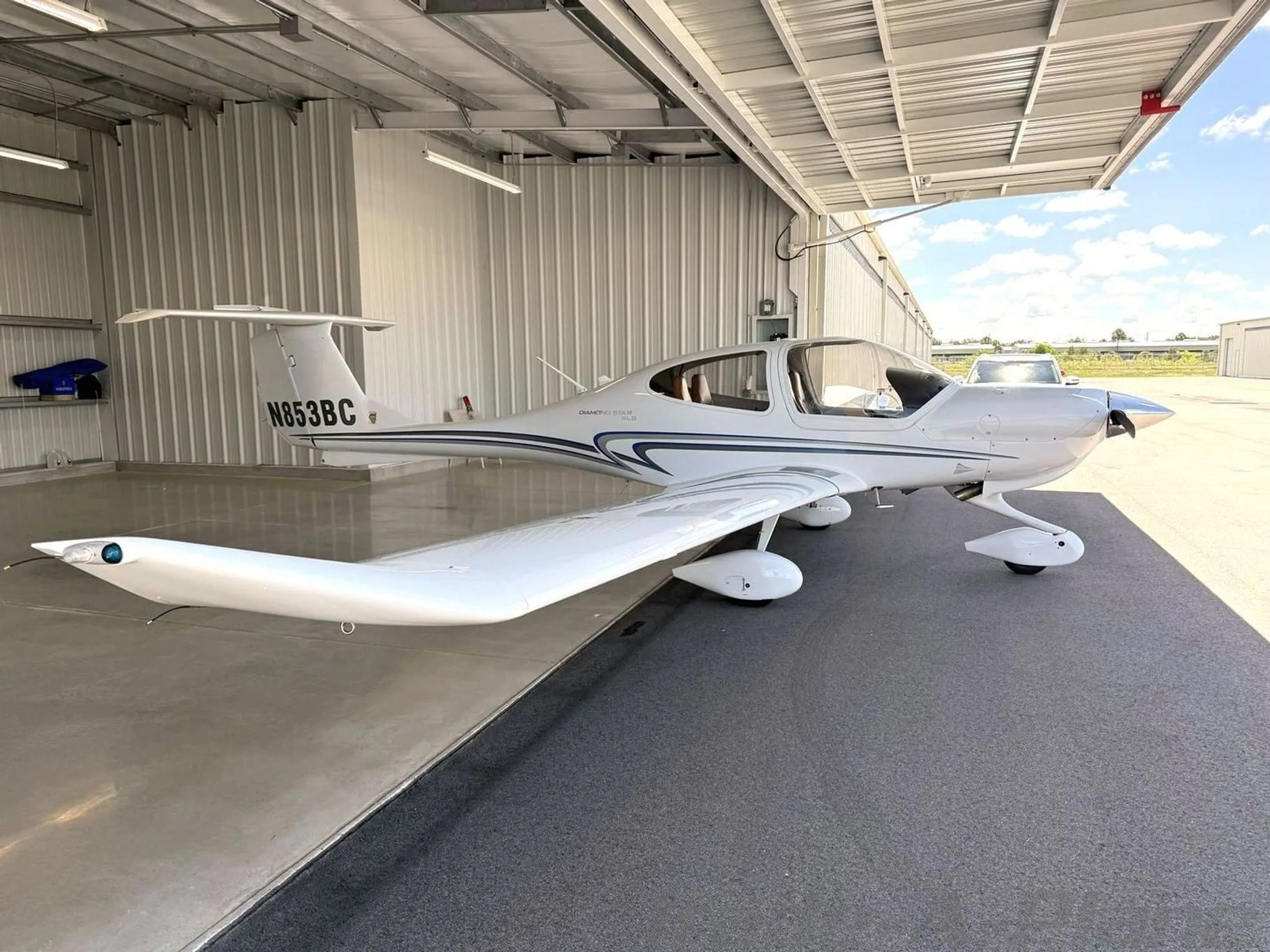 A side view of a white Diamond Star DA40 XLS aircraft with blue and grey stripes, tail number N853BC, parked partially in a hangar.
