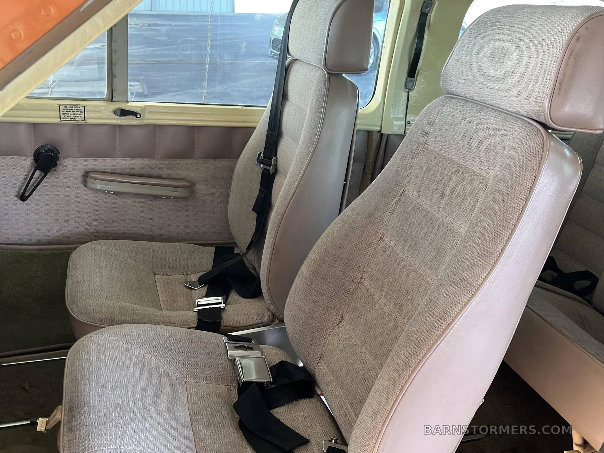 The interior cabin of an aircraft showing two rows of light brown fabric seats with seatbelts.