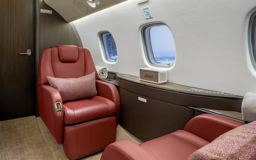 A close-up of a red leather reclining seat next to a window in a private jet cabin, with a Marshall speaker on the side table.