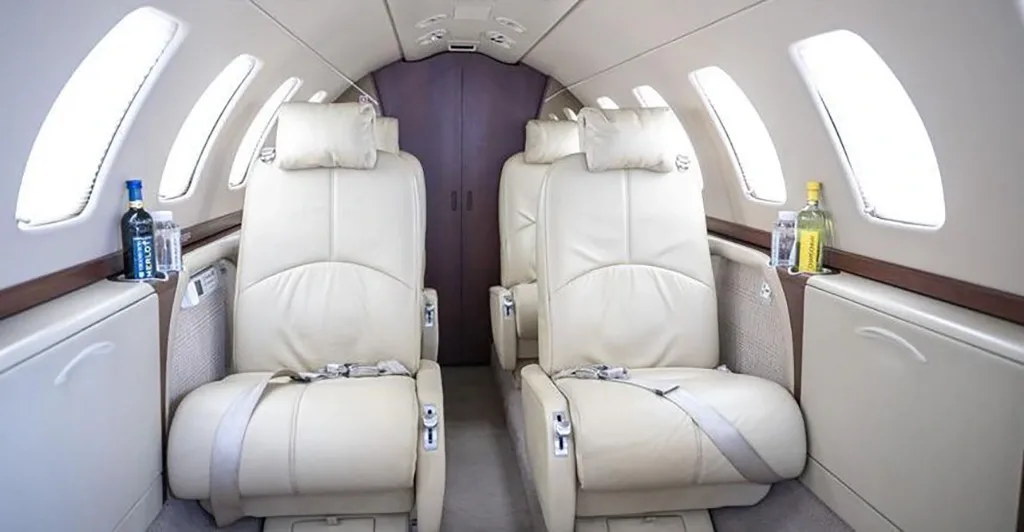 The interior cabin of a private jet with two rows of light beige leather seats facing each other, and bottles on side tables.