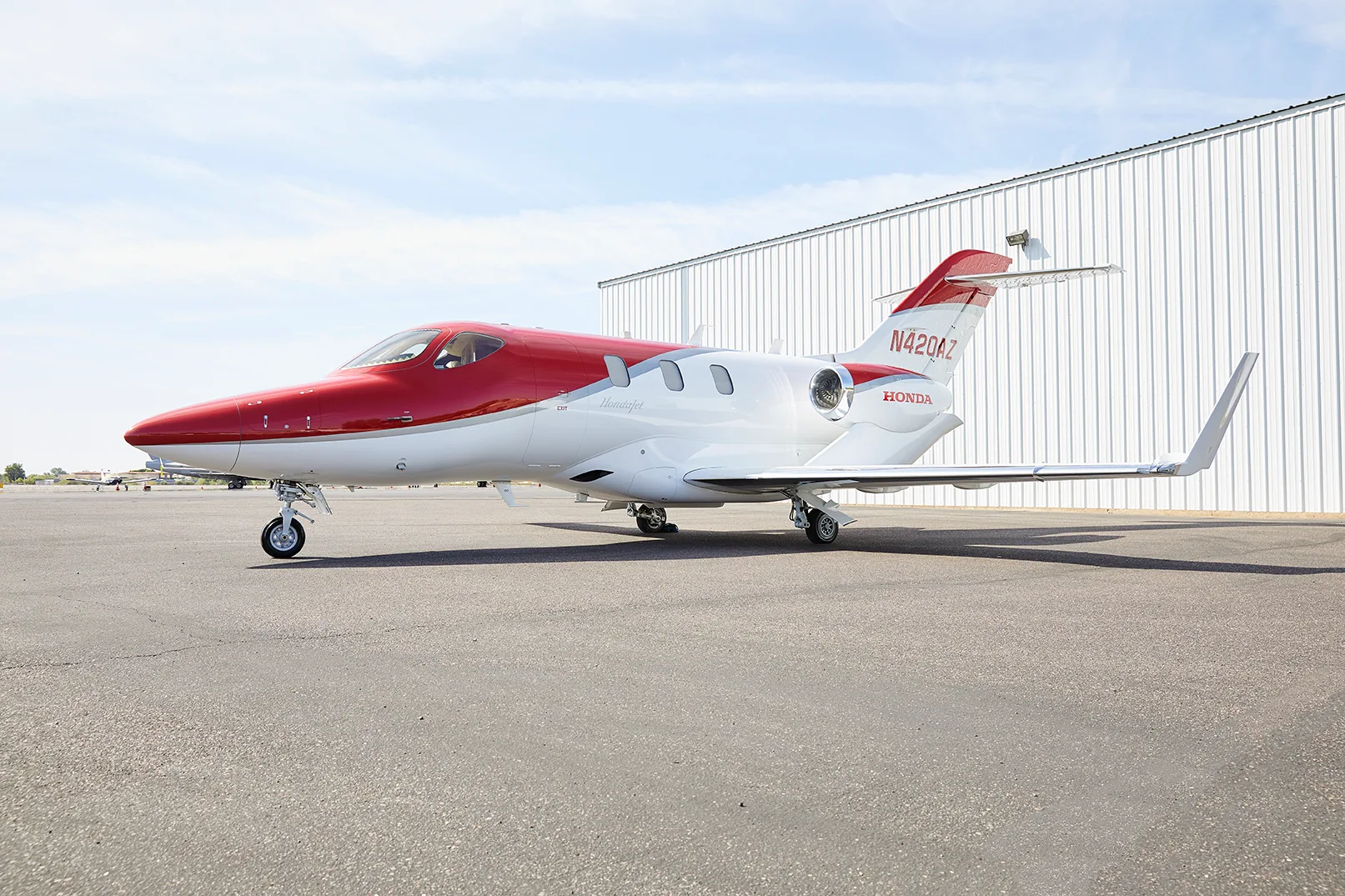 A red and white HondaJet HA-420 aircraft, registration N420AZ, is parked on an asphalt tarmac in front of a white hangar.