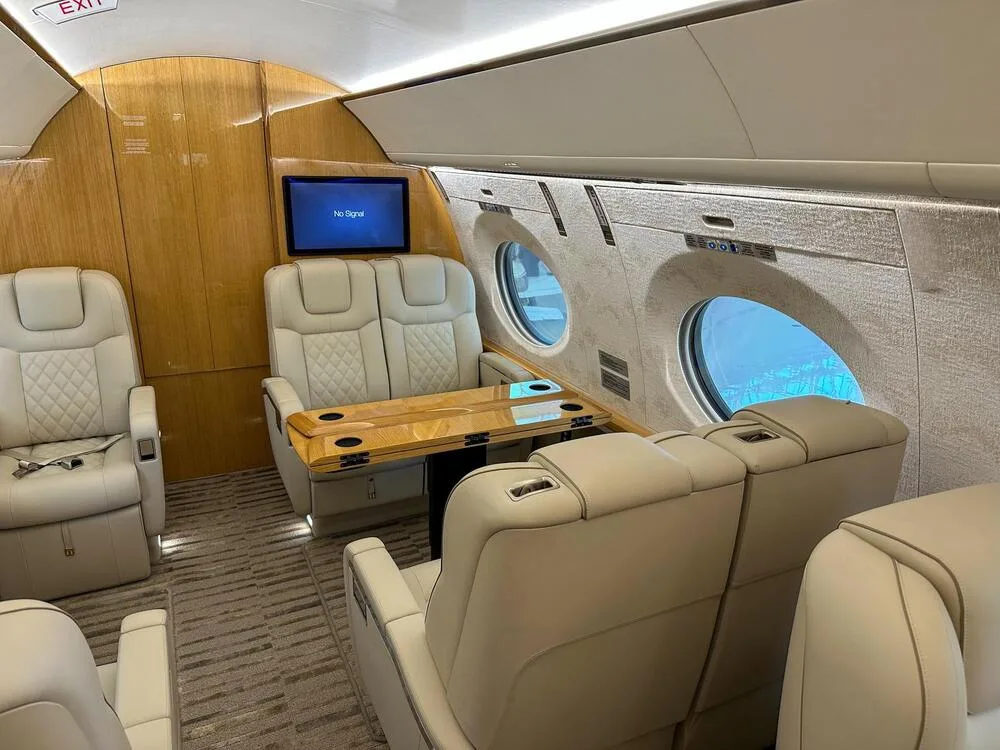 A view inside a private jet cabin showing light-colored leather seats, a wooden table, and large oval windows.