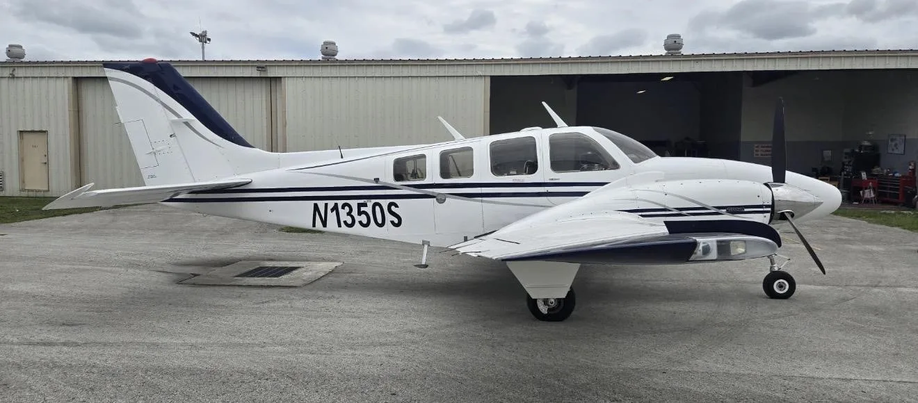 A white Beechcraft Baron N1350S with blue and grey stripes parked on a tarmac with a hangar in the background.