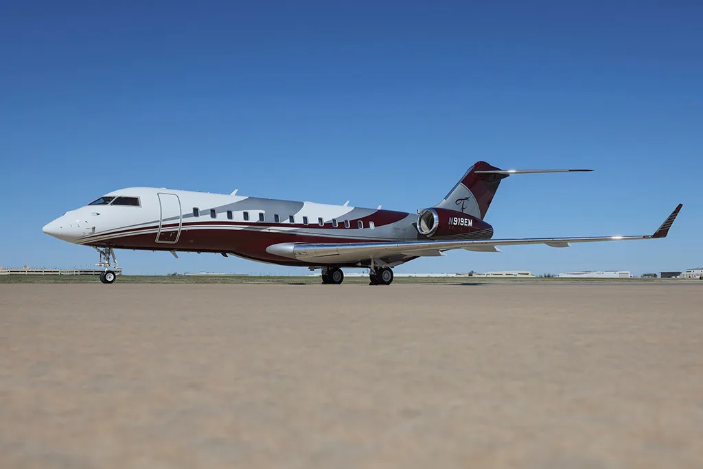 A Bombardier Global Express business jet with a white and maroon paint scheme, registration N919EM, parked on a tarmac under a clear blue sky.