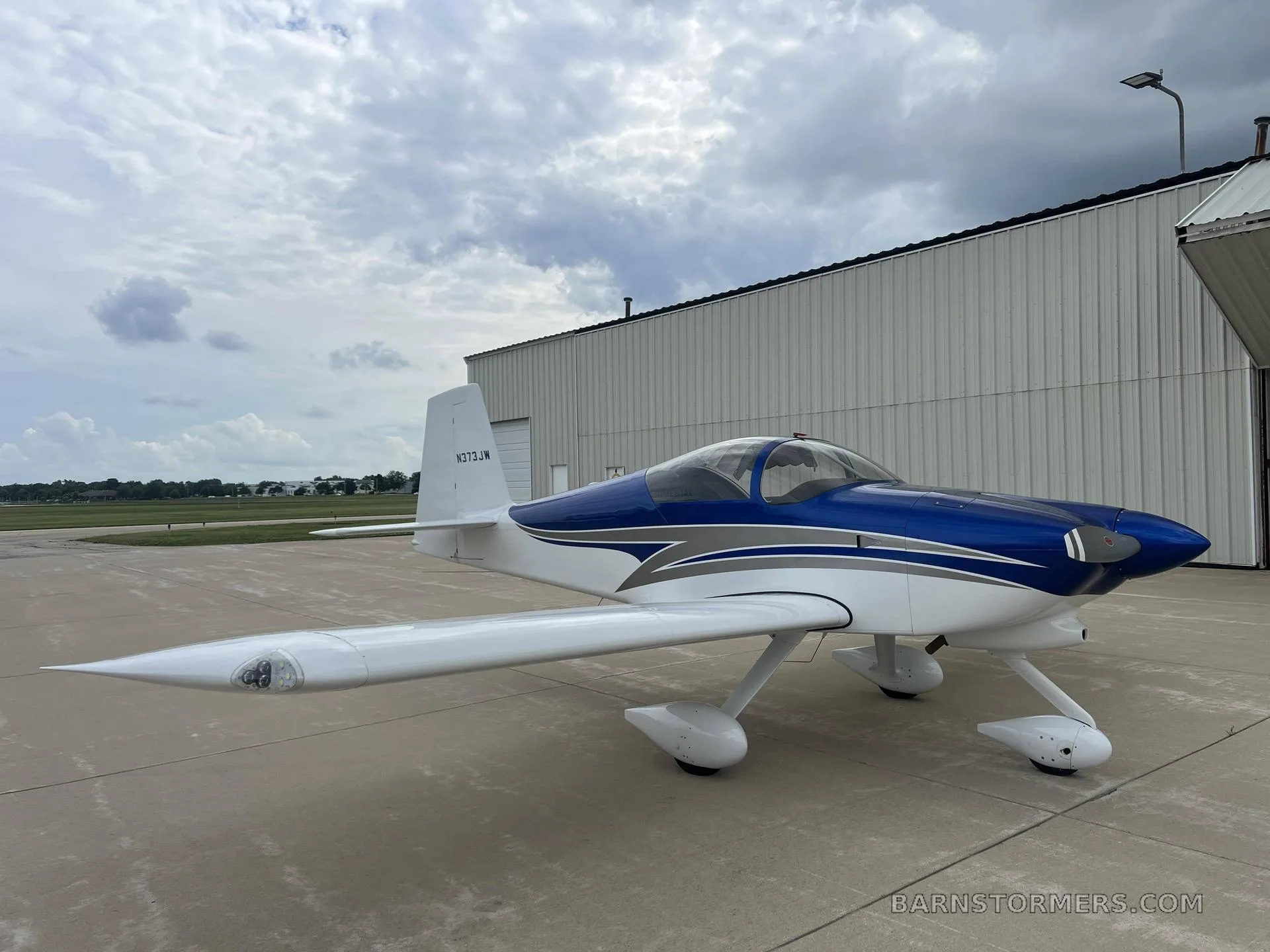 A white and blue single-engine aircraft, N373JW, with a bubble canopy, parked on a concrete tarmac next to a hangar under a cloudy sky.