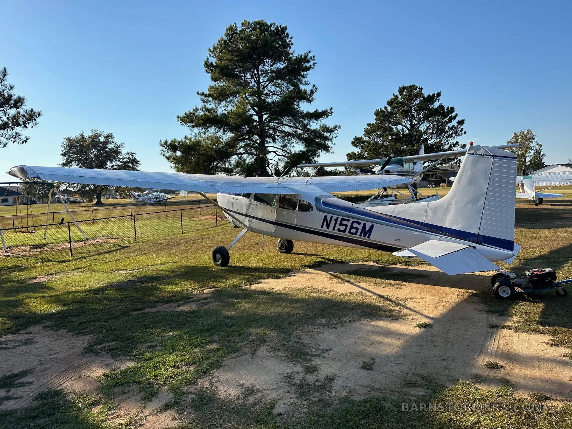 A white and blue Cessna 180 aircraft parked on a sunny grassy field next to a fence and trees.