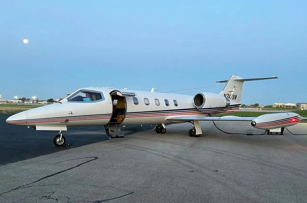 A white Learjet with red and blue stripes, registration N747BW, parked on an airfield at dusk with its door open.