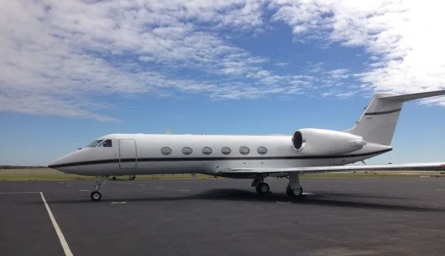 A white Gulfstream business jet with black trim is parked on an airport tarmac under a partly cloudy blue sky.
