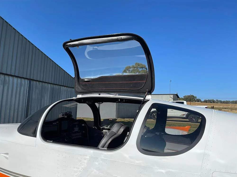 The side of a white aircraft with an open canopy reveals the dark interior and part of the instrument panel.